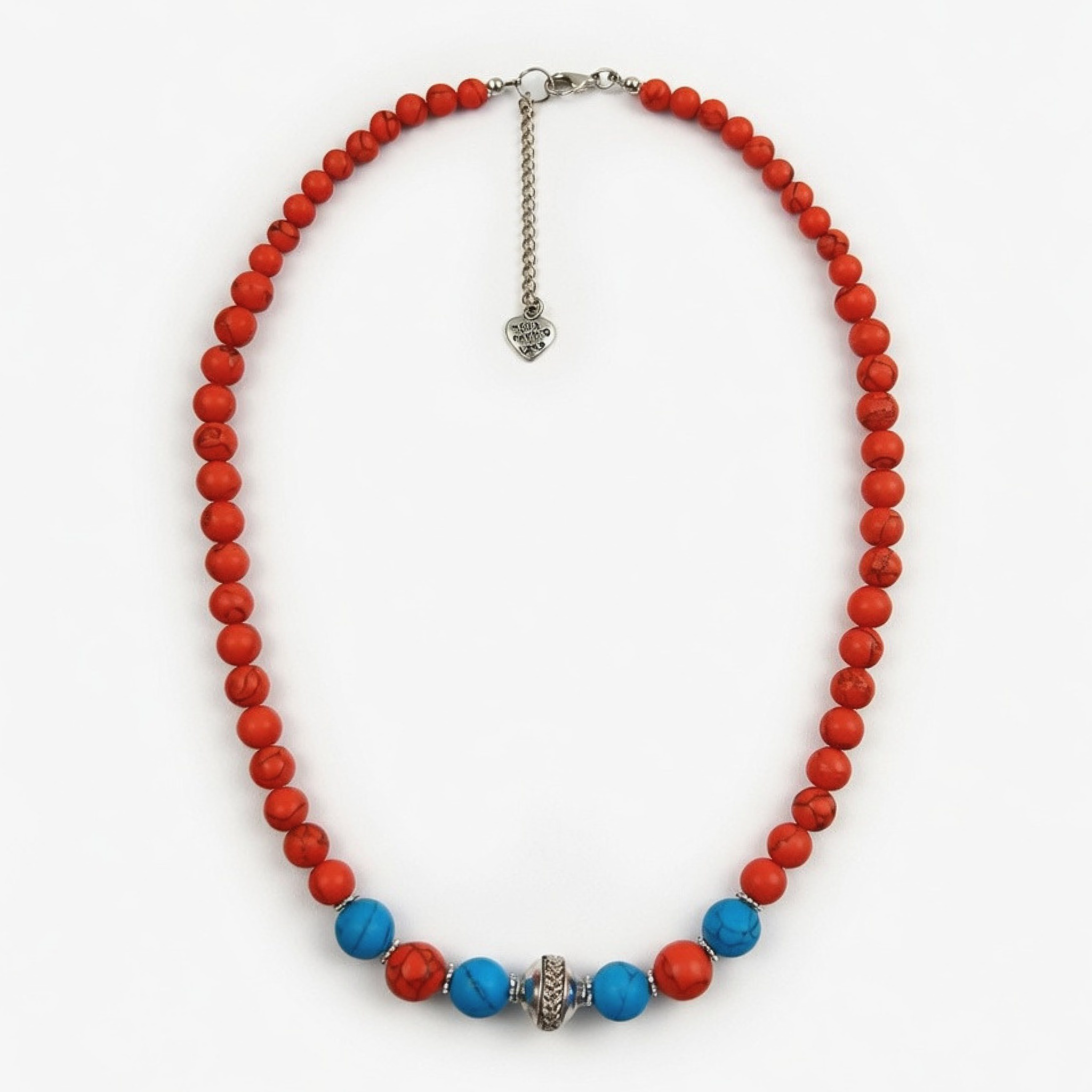 Eine Kette aus roten Howlith-Perlen, im unteren Bereich akzentuiert mit blauen Türkis-Perlen und einem gemusterten Silberelement in der Mitte, flach liegend vor weißem Hintergrund.
