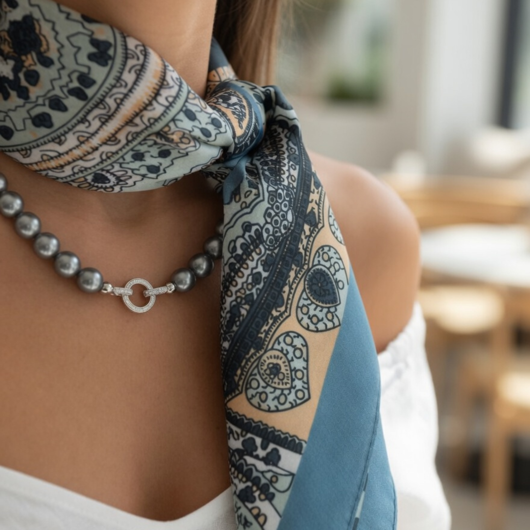 Nahaufnahme eines seitlich geknoteten Paisley-Halstuchs in Taubenblau und Mint, kombiniert mit einer anthrazitfarbenen Perlenkette. Das Model trägt ein schulterfreies weißes Top, wodurch das hochwertige Accessoire im Fokus steht.