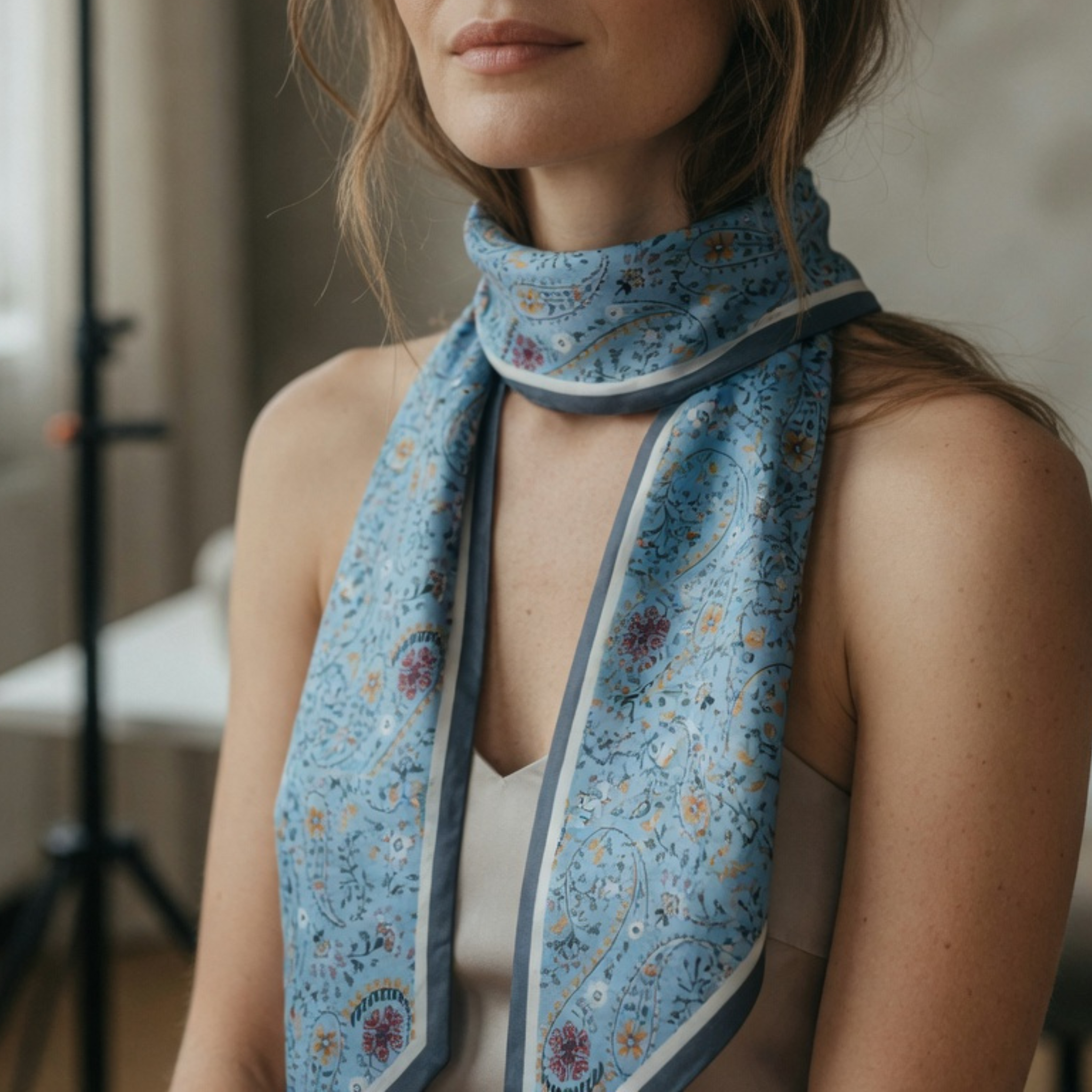 Fokus auf ein hellblaues Twilly-Halstuch mit Paisley-Print, das eng um den Hals geschlungen ist. Die herabhängenden Enden zeigen das filigrane Muster aus Blumen und Ranken vor dem Hintergrund einer dezenten Zimmerkulisse.