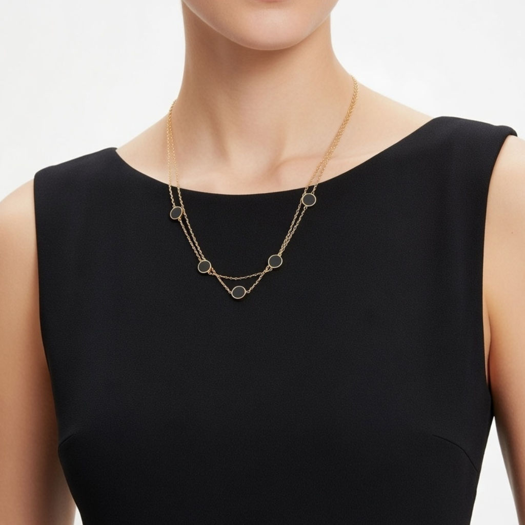 "Nahaufnahme einer Frau in einem ärmellosen schwarzen Kleid, die eine zweireihige goldene Halskette trägt. Die Kette ist in regelmäßigen Abständen mit runden, flachen schwarzen Plättchen besetzt, die harmonisch auf dem Dekolleté liegen."