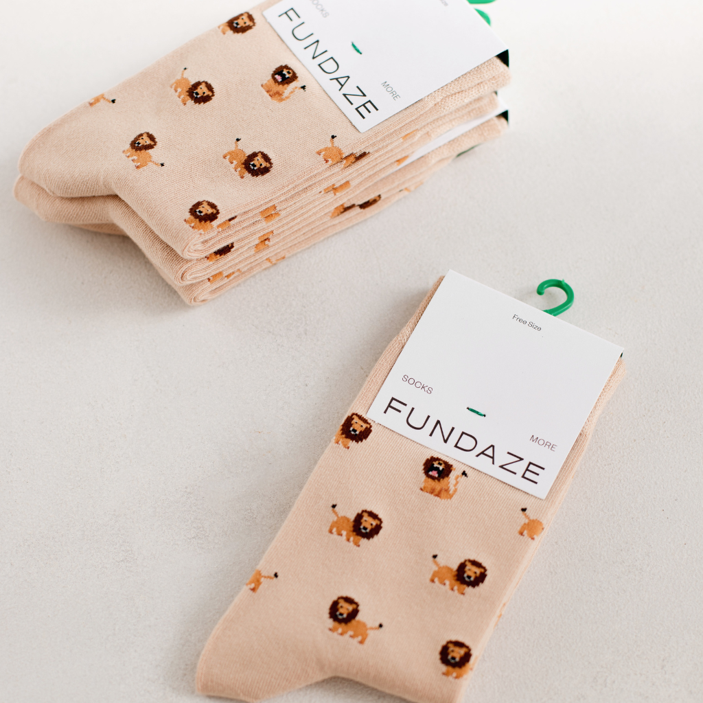 Mehrere Paar beige Socken der Marke FUNDAZE, ordentlich gestapelt und mit Etikett versehen. Die Socken zeigen ein flächendeckendes Muster aus kleinen, niedlichen Löwen-Illustrationen in Braun und Orange