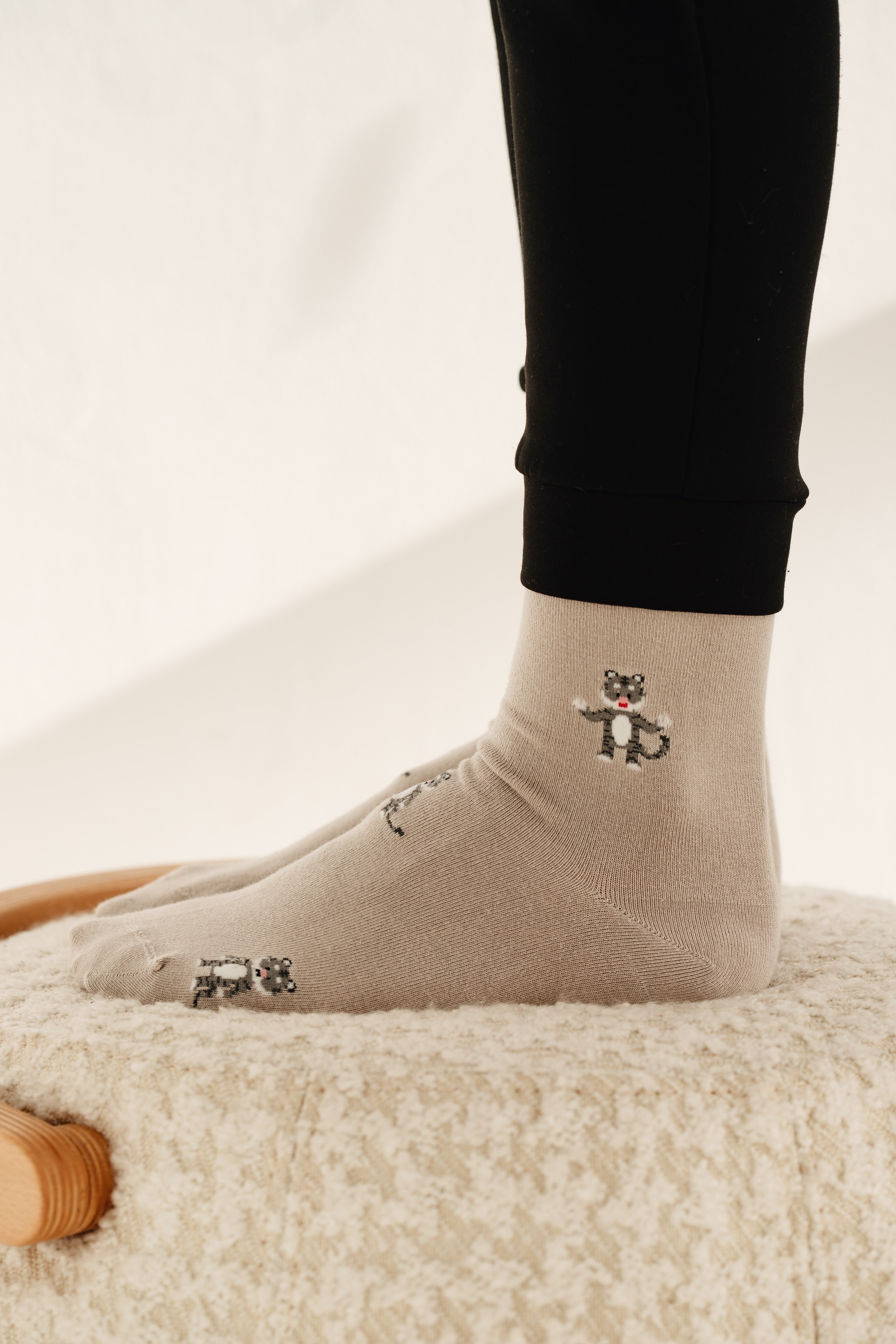Seitenansicht von zwei Füßen in beigen Socken mit kleinen Löwen-Prints, die auf einem hellen, weichen Teppich stehen.