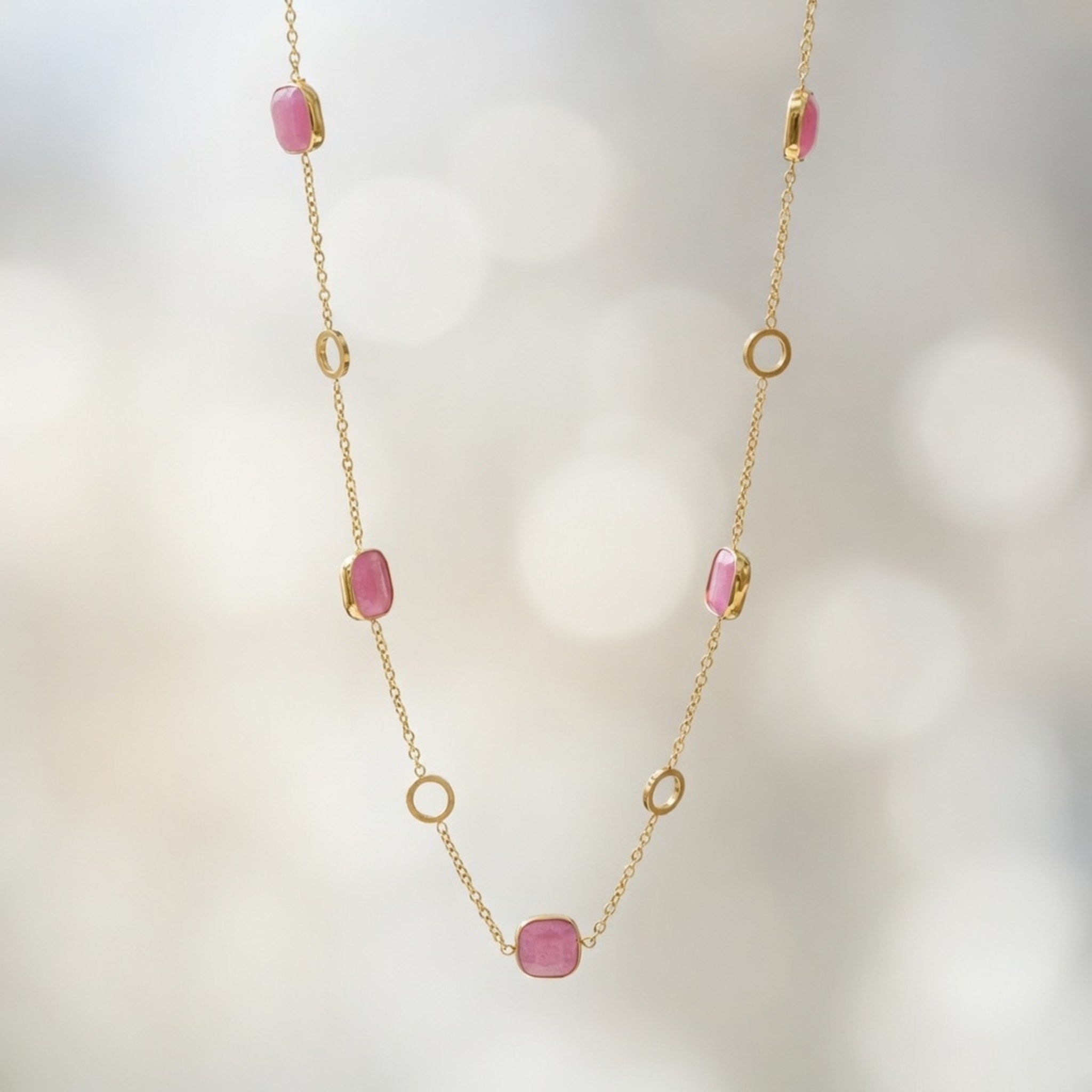Eine filigrane goldene Halskette, die vertikal vor einem hellen, unscharfen Hintergrund mit Bokeh-Effekt präsentiert wird. Die Kette ist abwechselnd mit quadratischen, rosa Edelsteinen in Goldfassung und offenen, kreisförmigen Goldelementen besetzt.