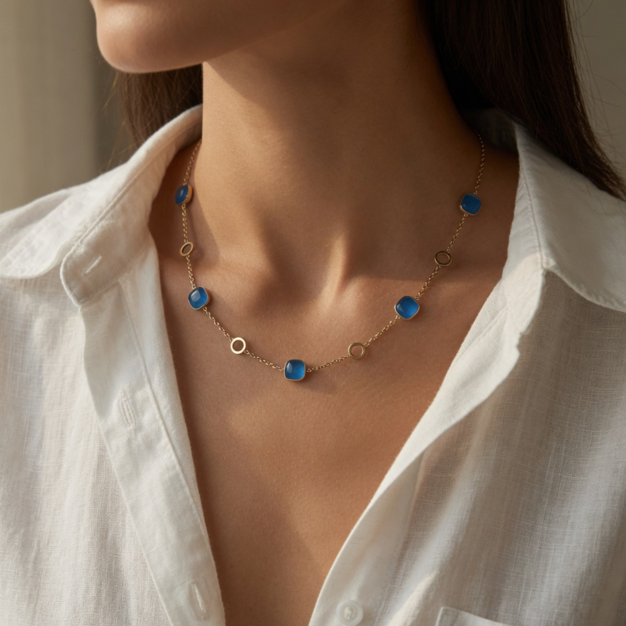 Nahaufnahme einer filigranen Goldkette am Hals einer Frau, besetzt mit fünf quadratischen, tiefblauen Steinen in Cabochon-Schliff, die abwechselnd mit kleinen, offenen Goldringen an einer feinen Ankerkette befestigt sind.