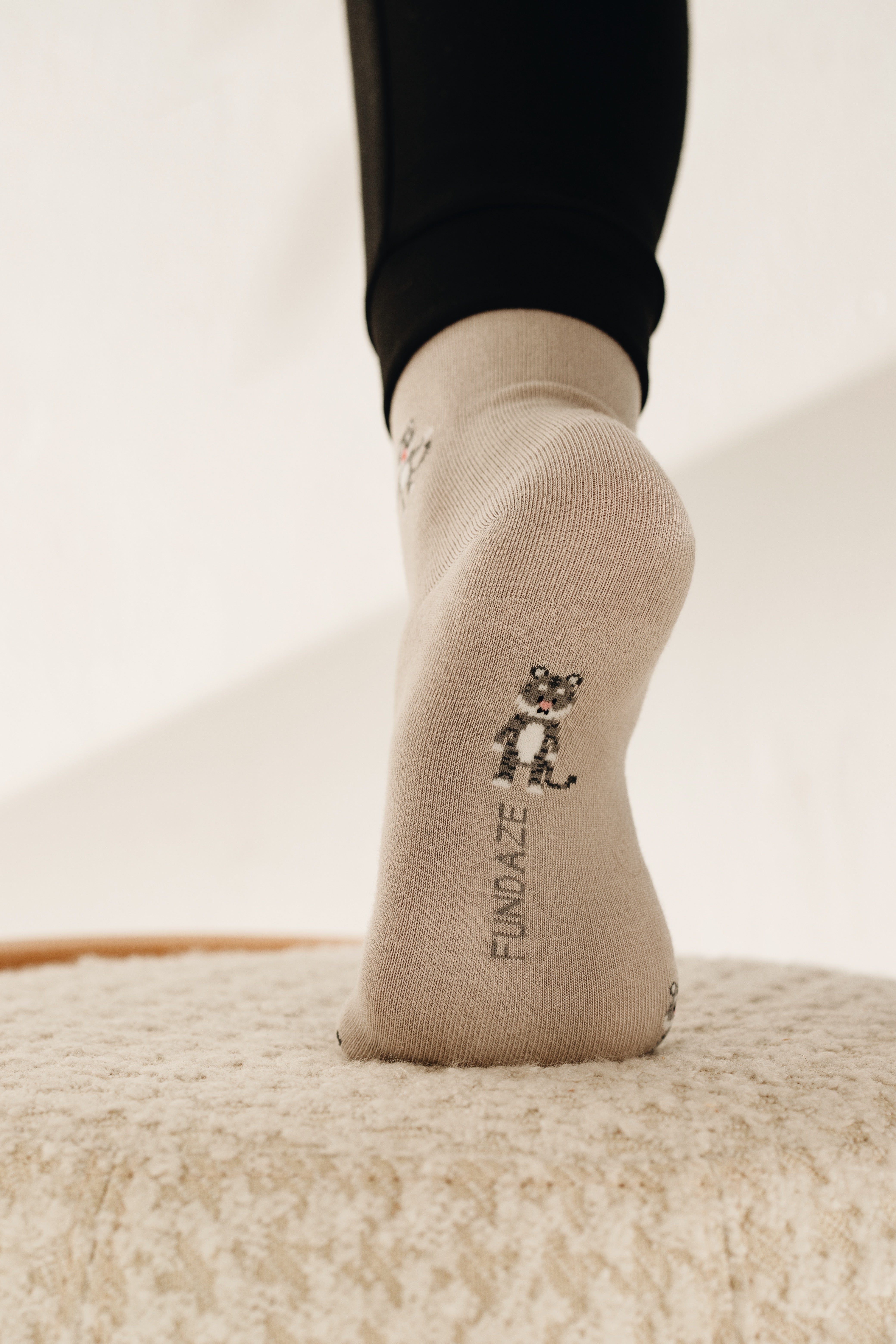 Rückansicht einer beige-melierten Socke am Fuß, die eine kleine Illustration eines stehenden Tieres mit gestreiftem Schwanz sowie den vertikalen Markenschriftzug „FUNDAZE“ zeigt.