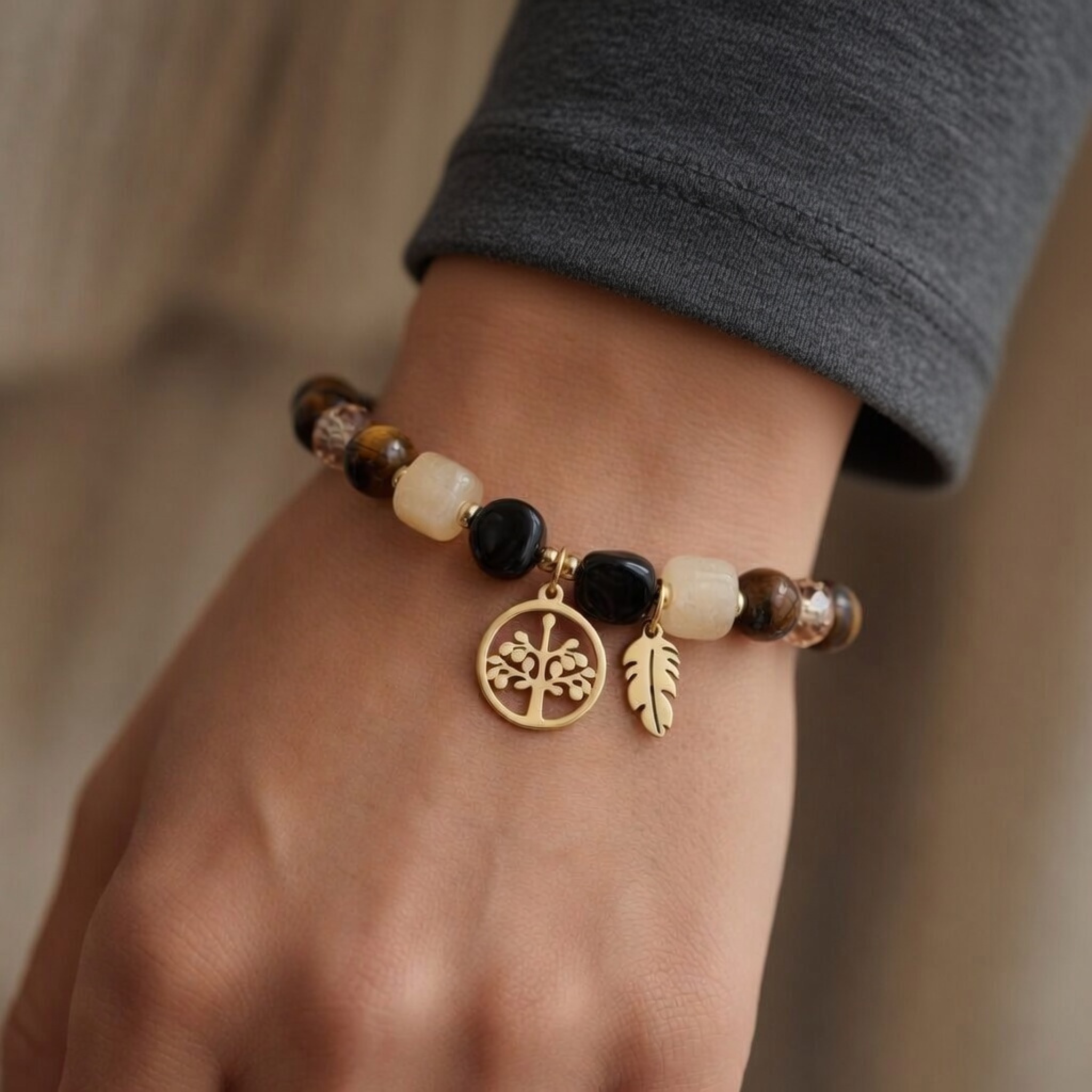 Damen-Perlenarmband am Handgelenk mit braunen Tigerauge-Steinen, schwarzen und beigen Perlen. Zwei goldene Anhänger zeigen einen filigranen Lebensbaum im Kreis und eine kleine Feder. Elegantes Boho-Schmuckstück kombiniert mit einem grauen Langarmshirt.