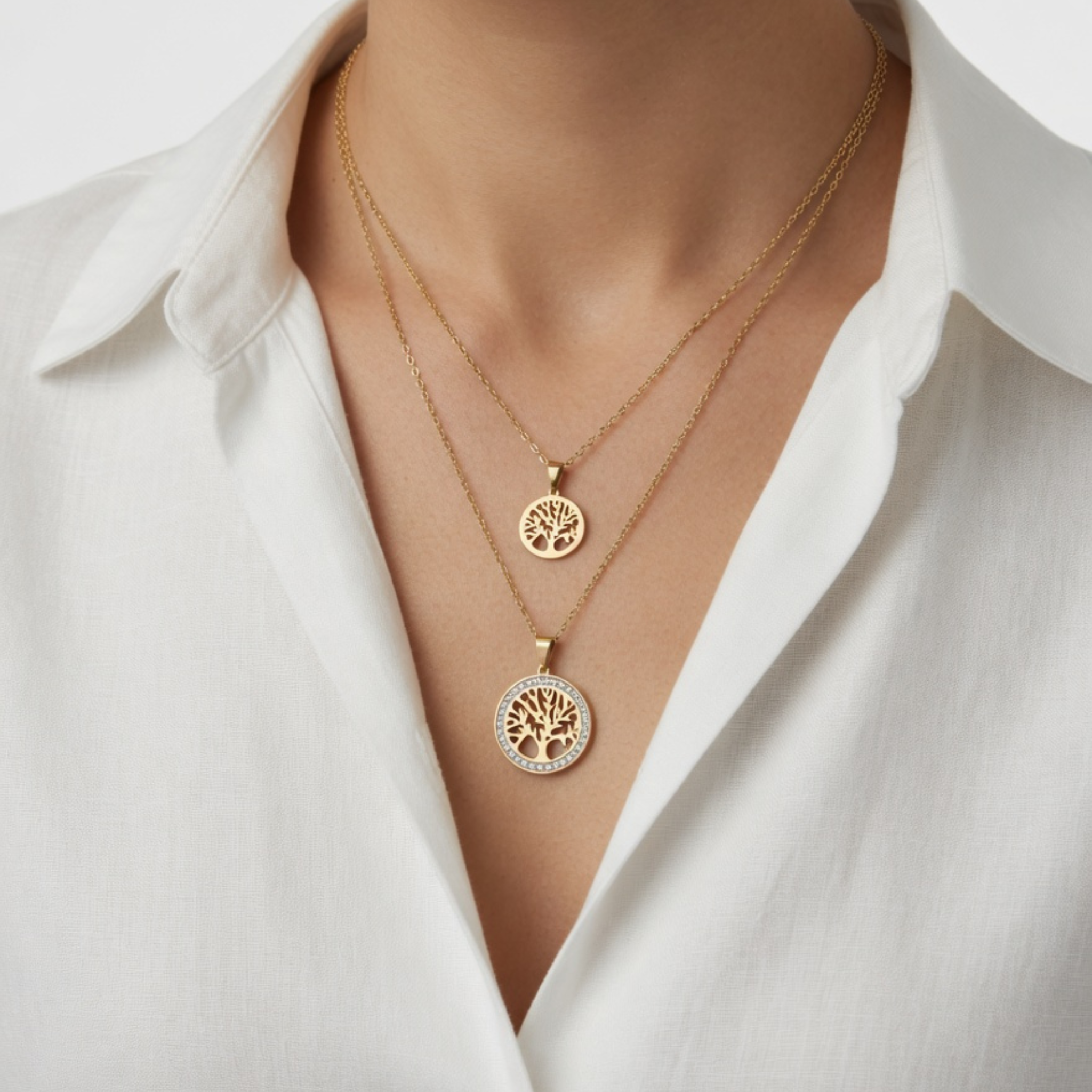 Eine Frau trägt zwei verschieden lange, goldene Halsketten mit runden Lebensbaum-Anhängern zu einer eleganten weißen Bluse im Layering-Stil.