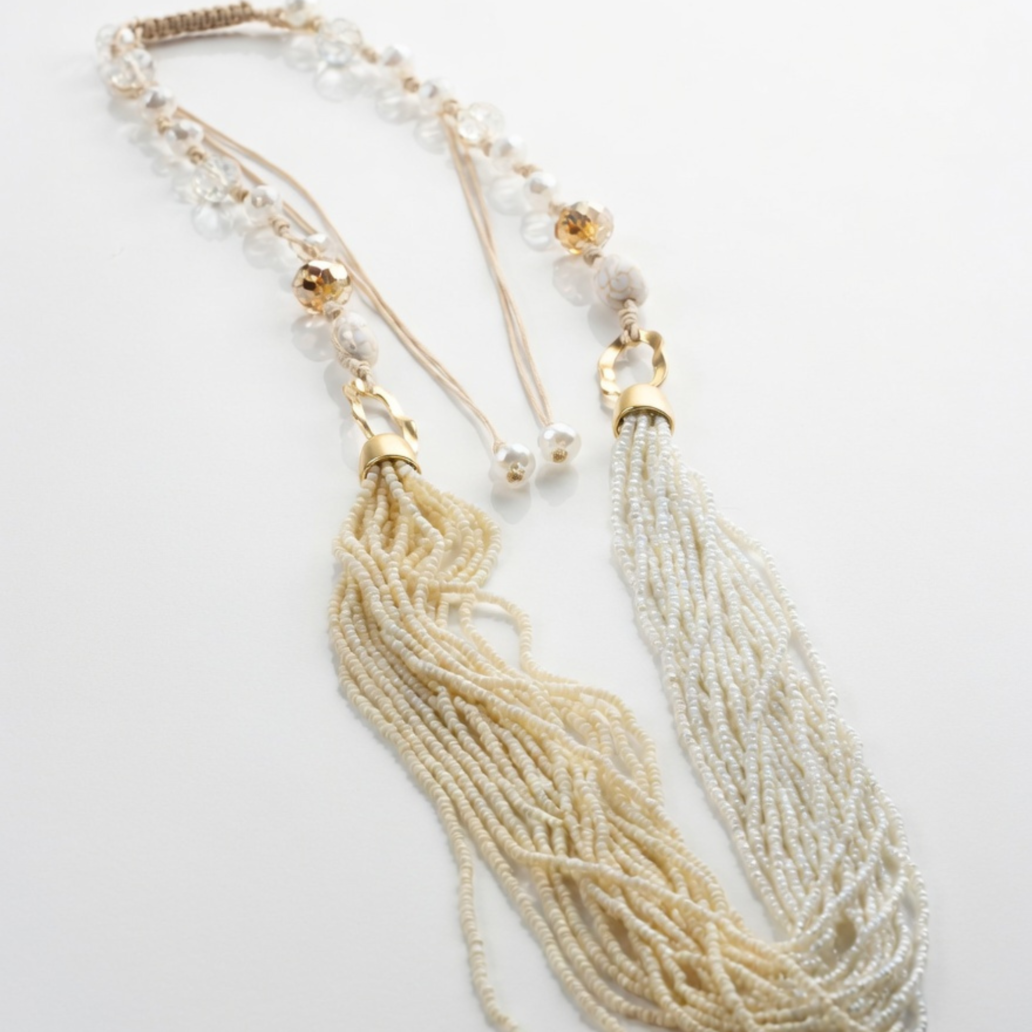 Produktfoto einer langen, zweigeteilten Statement-Kette auf hellem, leicht reflektierendem Untergrund. Zu sehen sind dichte Perlenstränge in Creme und Reinweiß, geschliffene Glasperlen am Band sowie eine Kordel zum Binden.