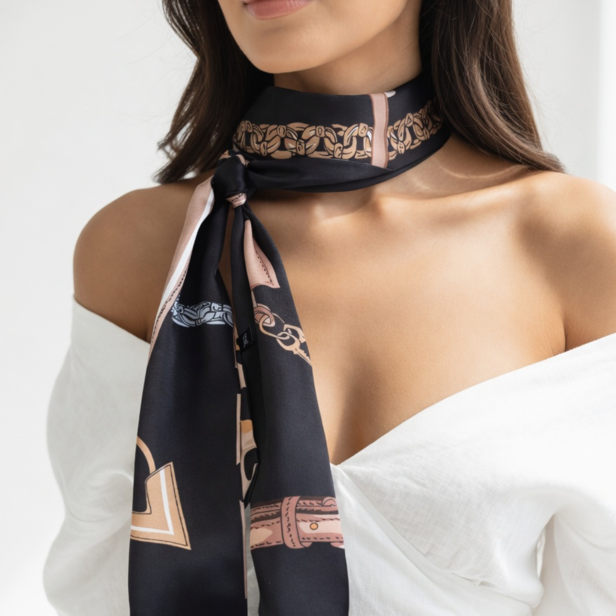 Das schmale Bandana ist eng um den Hals eines Models geknotet, wobei die schwarze Seite mit den goldenen Kettenelementen dominiert. Die langen Enden hängen locker über ein schulterfreies weißes Top und betonen den eleganten Kontrast des Accessoires.