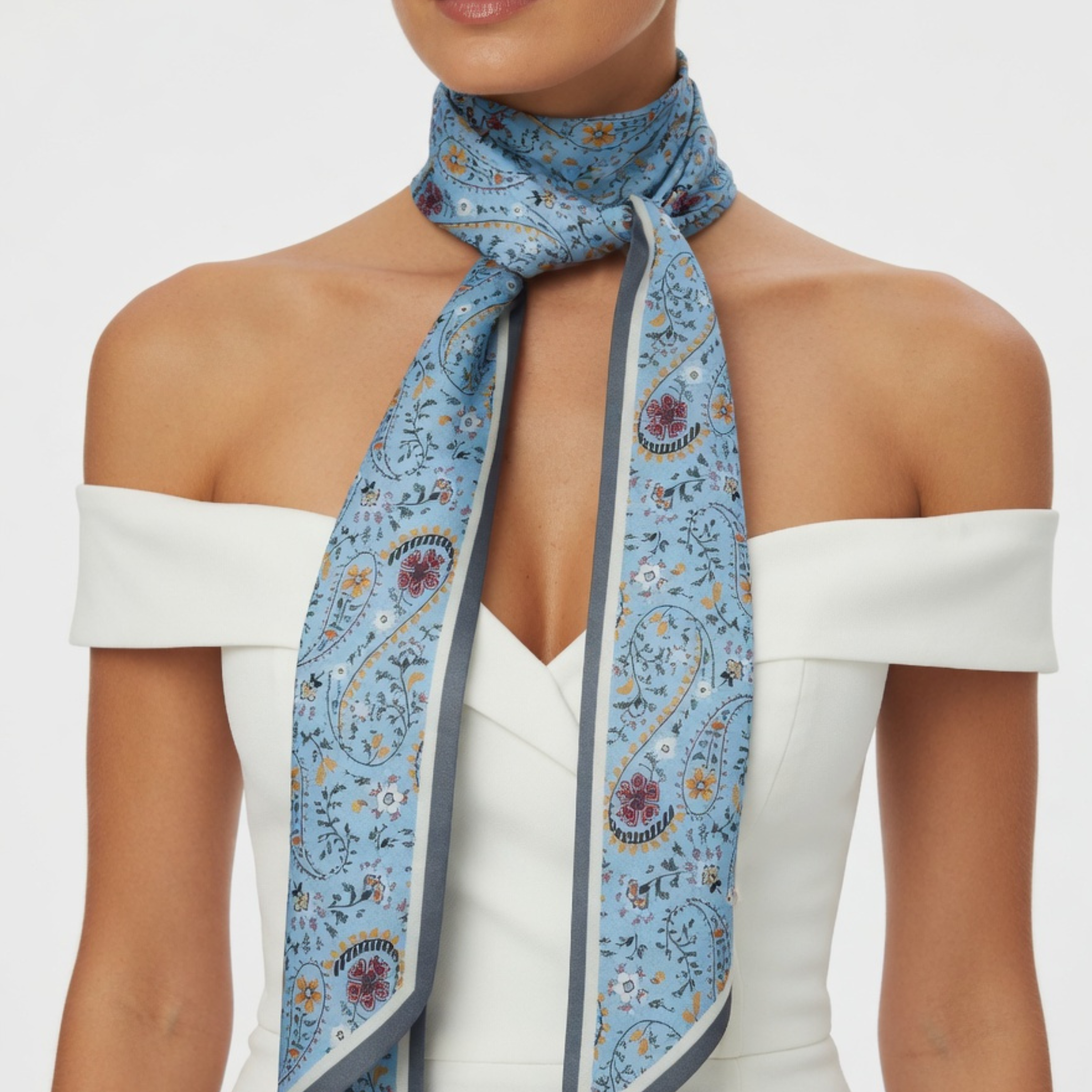 Nahaufnahme eines Models in einem weißen, schulterfreien Kleid, das ein schmales hellblaues Halstuch trägt. Das Tuch ist vorne mit einem einfachen Knoten gebunden, wodurch das detailreiche Paisley-Muster und die spitz zulaufenden Enden besonders zur Geltung kommen.