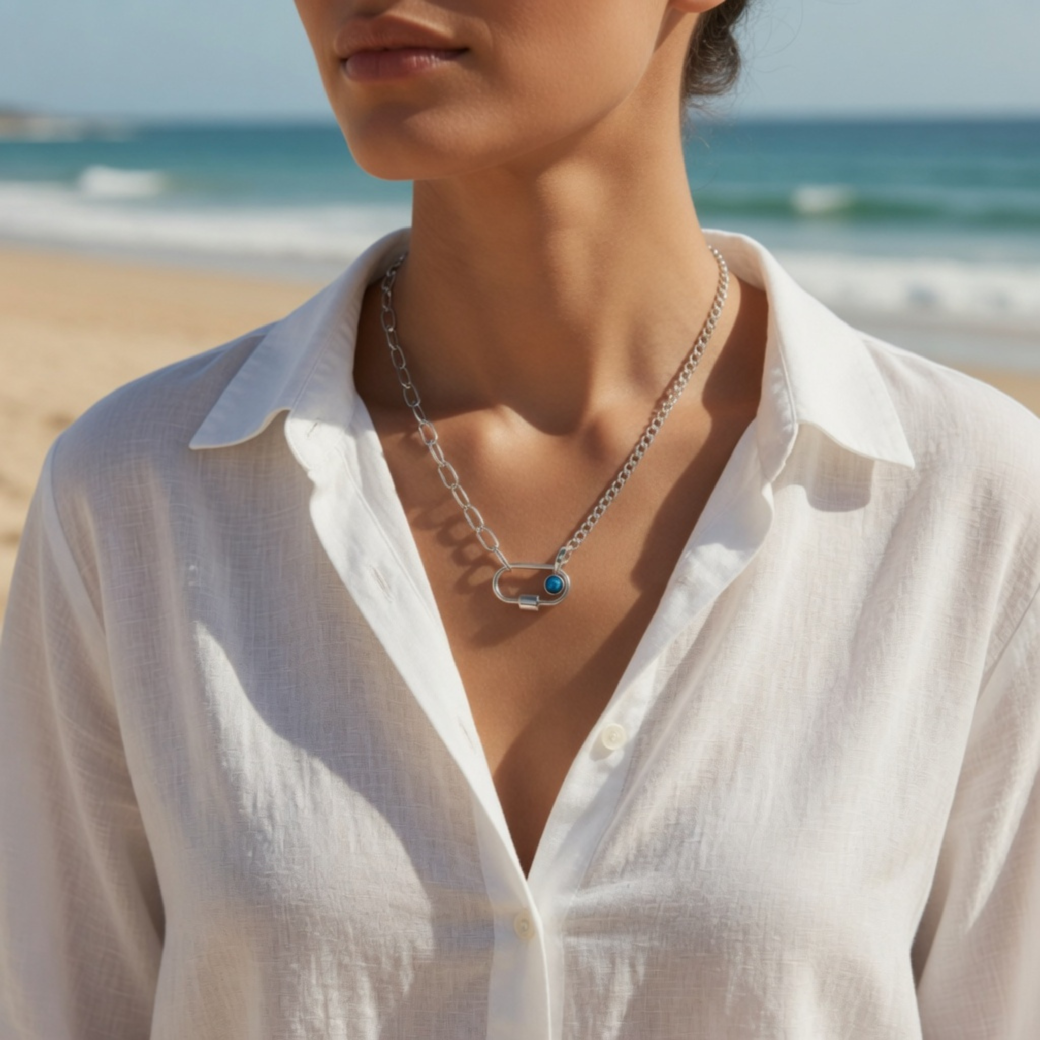 Eine Frau am Strand im weißen, leicht geöffneten Leinenhemd, kombiniert mit einer silbernen Gliederkette und einem Karabiner-Verschluss samt blauem Stein.