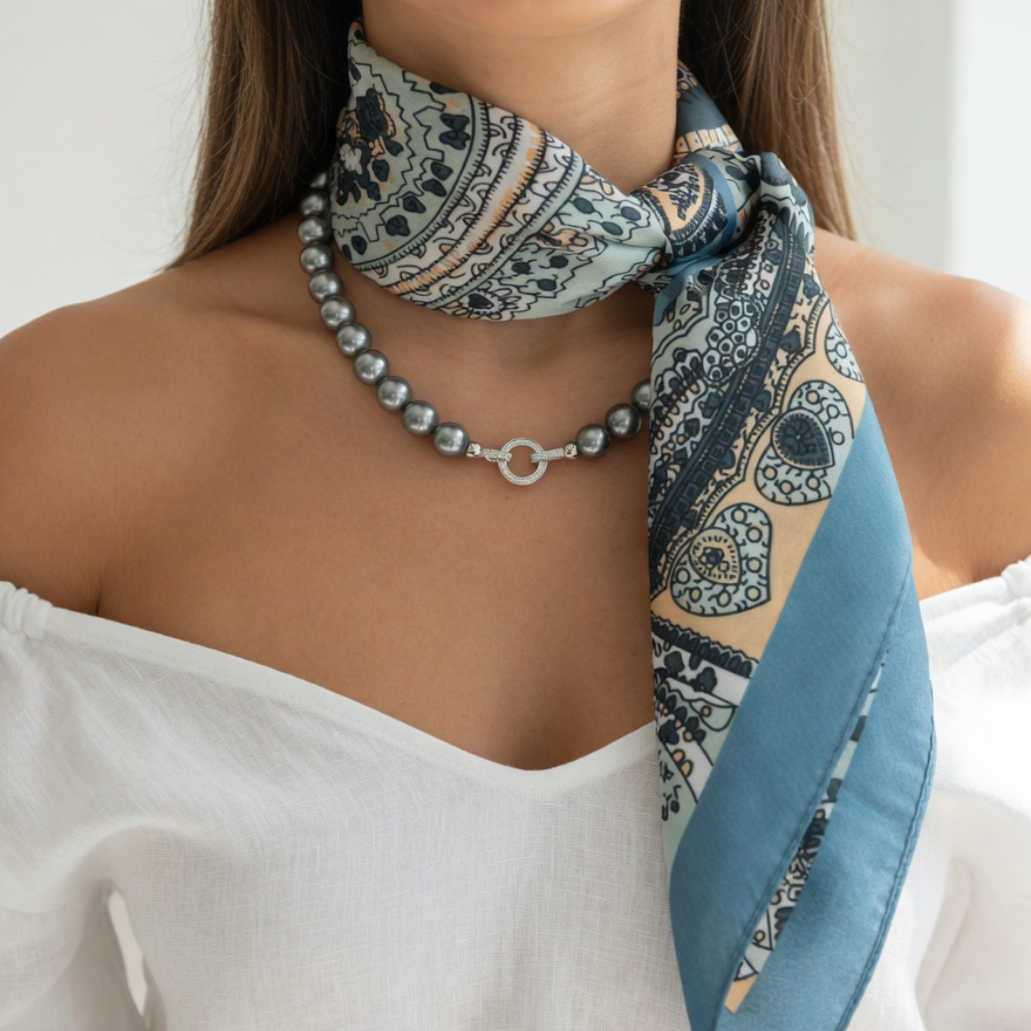Nahaufnahme eines seitlich geknoteten Seidentuchs mit Paisley-Muster in Taubenblau, Mint und Ocker. Das Model kombiniert das Accessoire mit einer anthrazitfarbenen Perlenkette mit silbernem Verschlussring zu einem schulterfreien weißen Top.