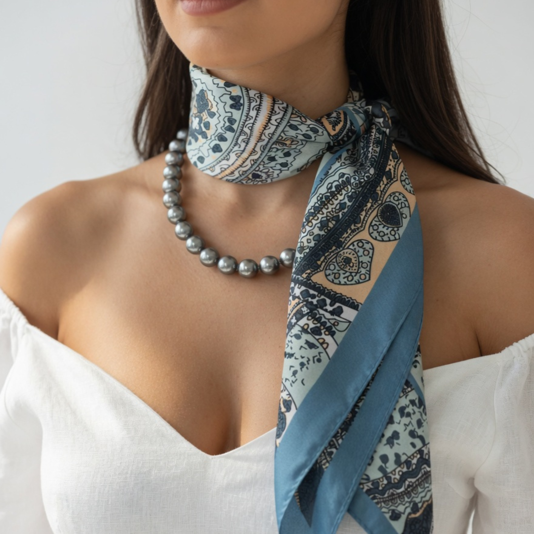 Ein am Hals geknotetes Damen-Halstuch mit Paisley-Muster in Mintgrün und Taubenblau, kombiniert mit einer anthrazitfarbenen Perlenkette. Das Tuch liegt elegant über der Schulter eines Models in einem weißen Off-Shoulder-Top vor neutralem Hintergrund.