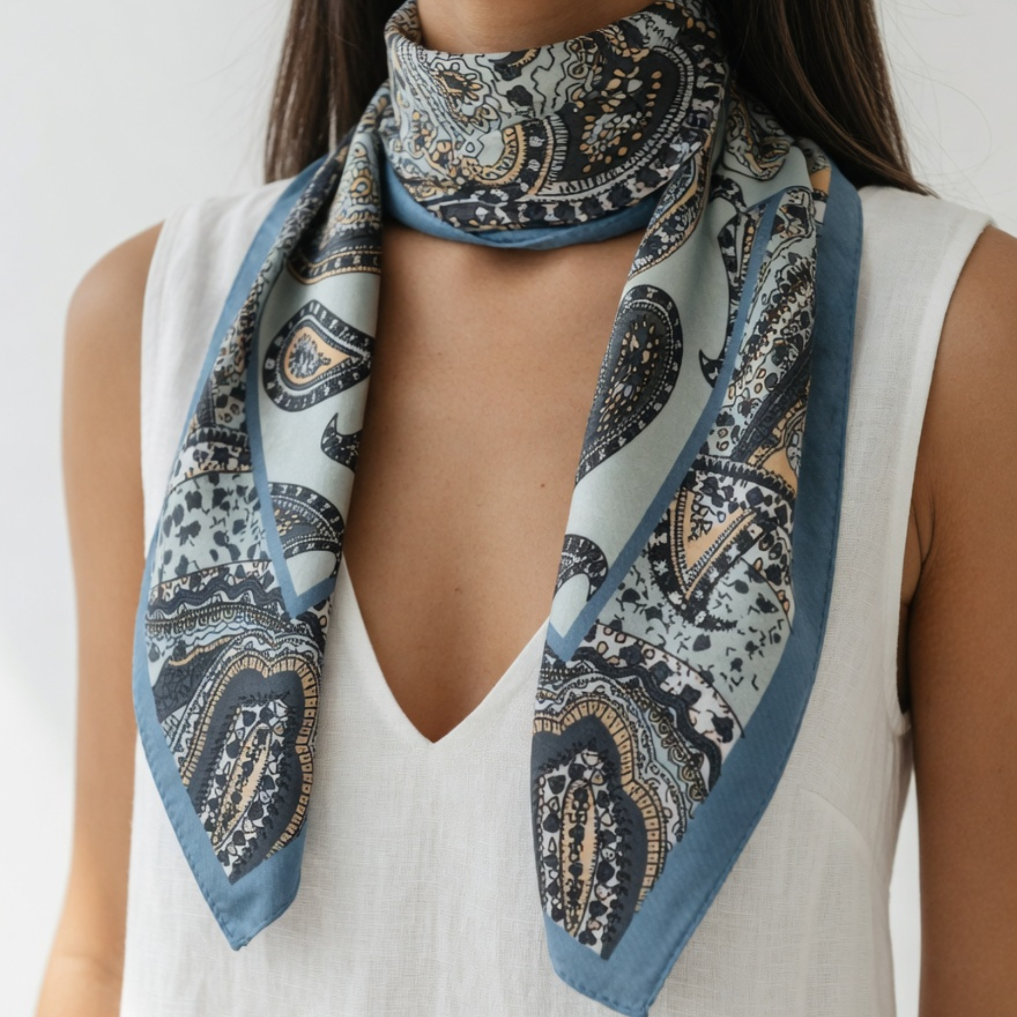 Das Paisley-Tuch liegt in einer lockeren Schlinge um den Hals, wobei die Enden symmetrisch über ein weißes ärmelloses Kleid fallen. Die feinen Ornamente und der blaue Kontrastrand betonen den sommerlich-eleganten Look.