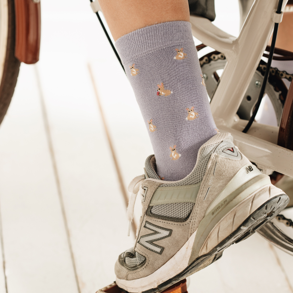 Modischer Lifestyle-Shot: Fliederfarbene Socken mit süßem Corgi-Hundemotiv, kombiniert mit sportlichen Sneakern beim Fahrradfahren. Ein perfektes Accessoire für einen lässigen Urban-Look.