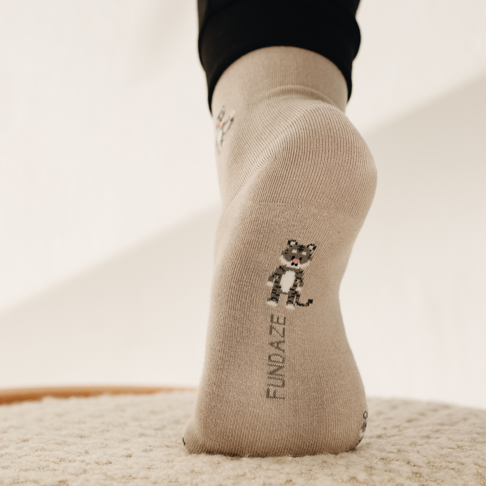 Rückansicht einer beige-melierten Socke am Fuß mit einer kleinen Löwen-Illustration über der Ferse und dem vertikalen Markenschriftzug „FUNDAZE“.