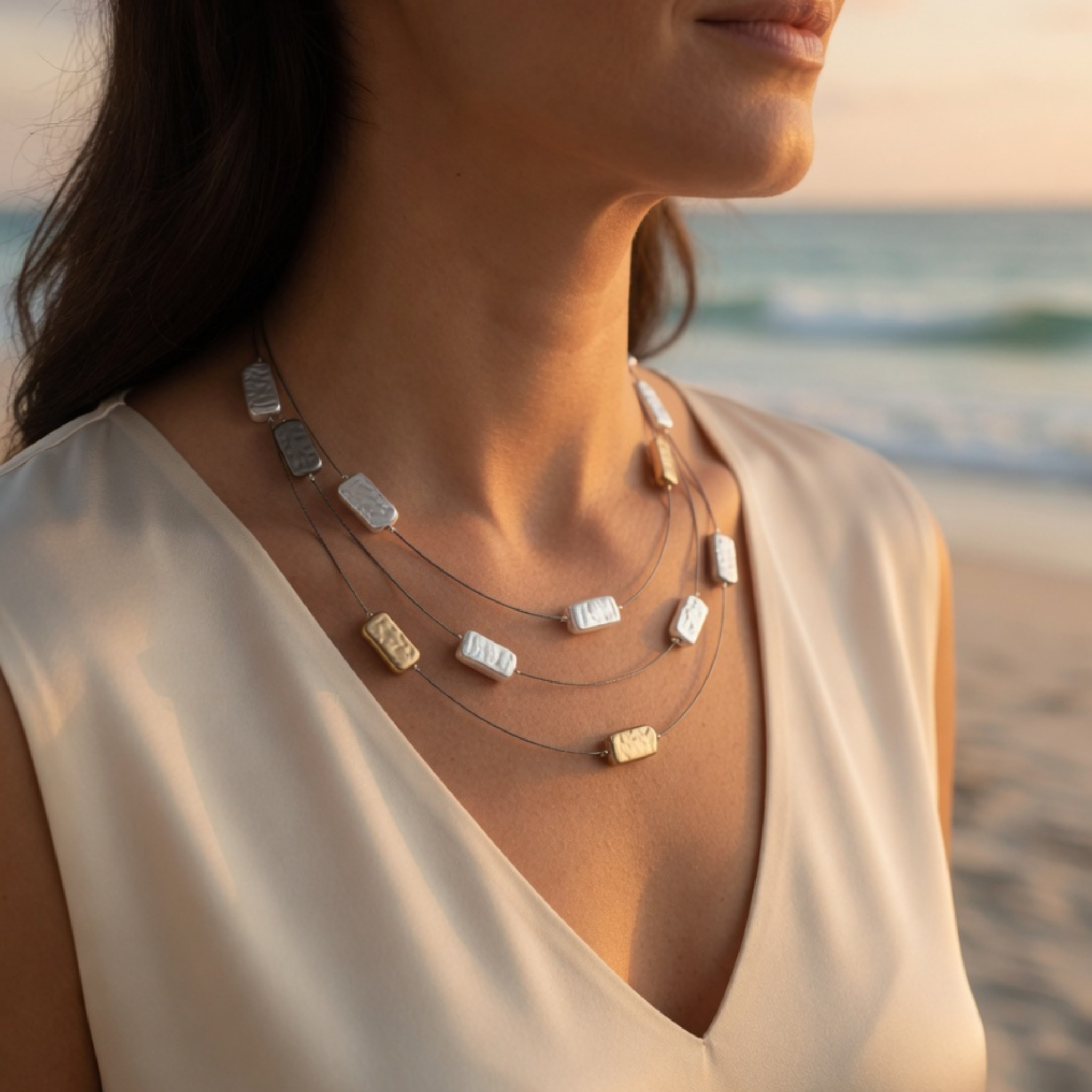 Nahaufnahme einer Frau am Strand im warmen Licht des Sonnenuntergangs, die eine feine, dreireihige Edelstahlkette mit silbernen und goldenen Schmuckplättchen trägt.