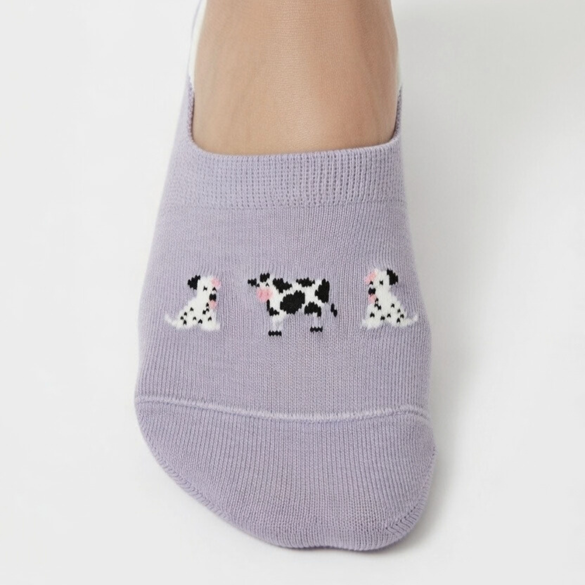 Nahaufnahme eines Fußes in einem fliederfarbenen Füßling. Auf dem Spann der Socke sind drei kleine, detaillierte Motive eingestickt: zwei weiße Hunde mit schwarzen Flecken (Dalmatiner-Stil) und eine schwarz-weiß gefleckte Kuh in der Mitte.