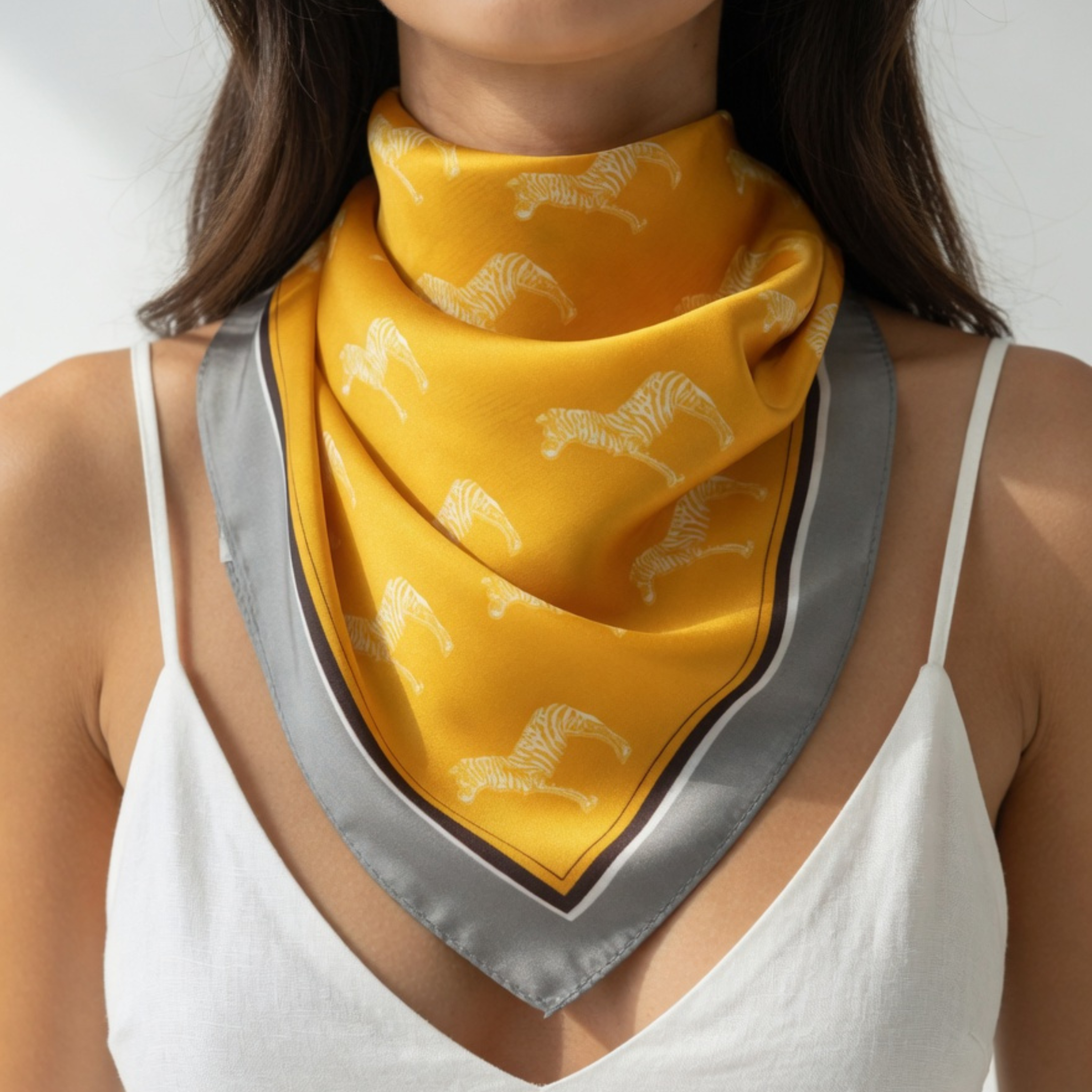 Ein senfgelbes Damen-Halstuch mit weißem Zebra-Muster, als Dreieckstuch über einem weißen Top getragen. Der graue Rand mit Kontraststreifen verleiht dem hochwertigen Accessoire einen modernen Safari-Look.