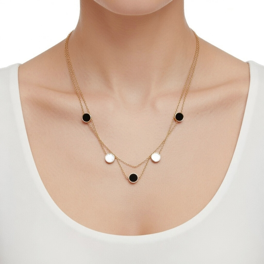 „Nahaufnahme einer Frau in einem weißen Oberteil, die eine zweireihige goldene Halskette trägt. Die Kette ist abwechselnd mit runden, flachen Plättchen in Schwarz und glänzendem Weiß (Perlmutt-Optik) besetzt, die in zwei Ebenen am Dekolleté hängen.“