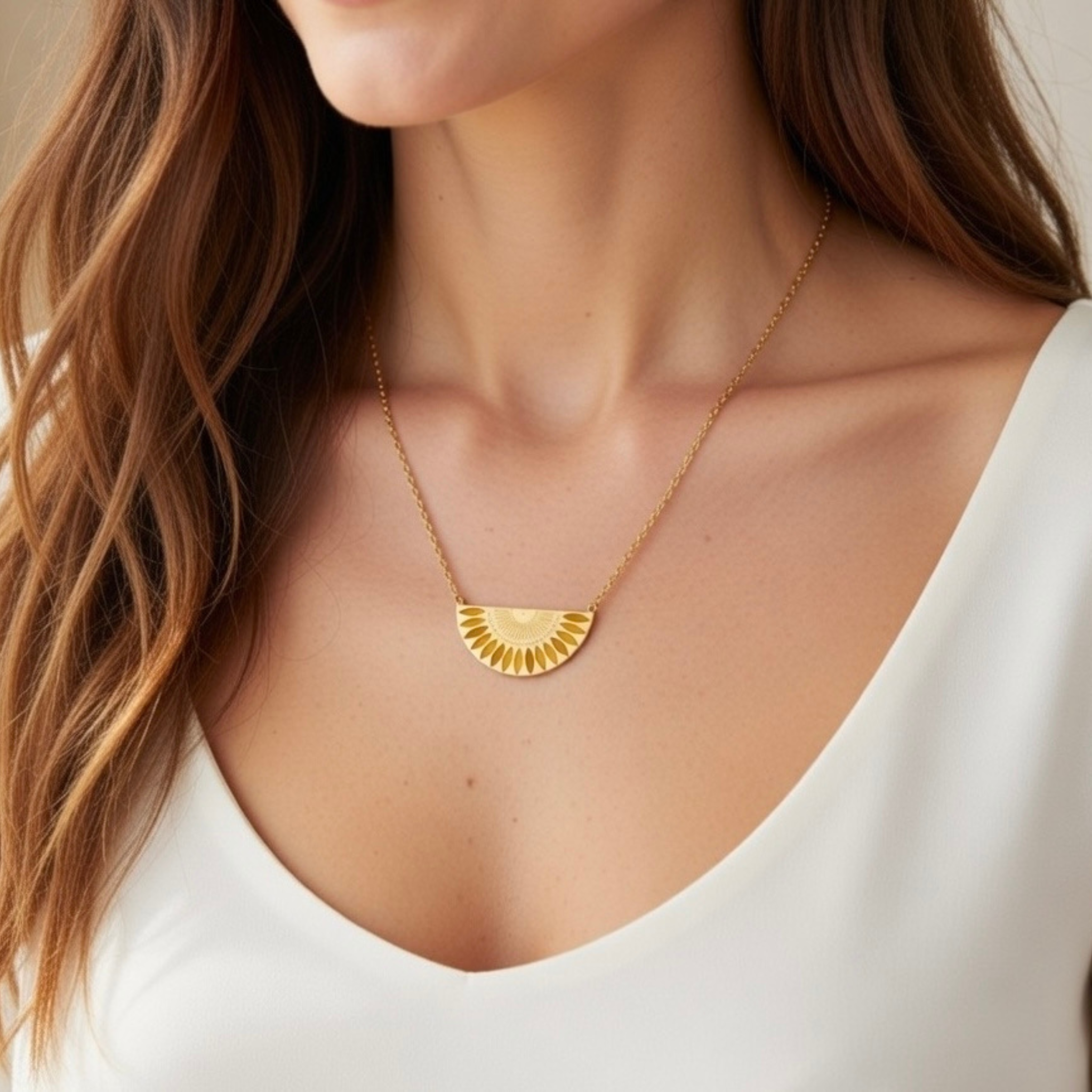 Nahaufnahme einer Frau mit einer goldenen Halskette. Der Anhänger hat die Form eines Halbkreises und ist mit einem filigranen Sonnenstrahlen-Muster graviert. Sie trägt dazu ein elegantes weißes Oberteil mit V-Ausschnitt.