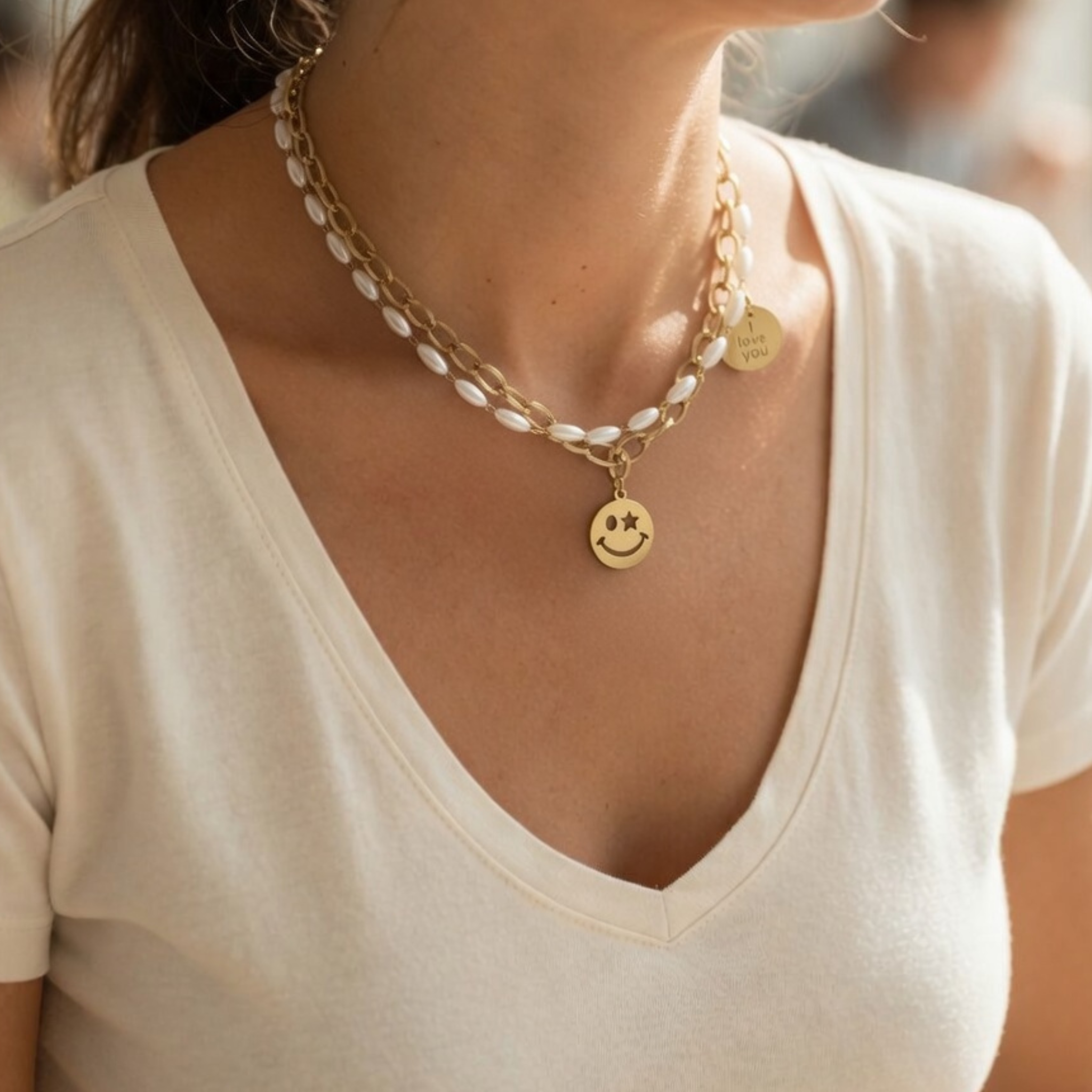 Nahaufnahme einer zweireihigen Halskette, getragen von einem Model. Die Kombination besteht aus einer goldenen Gliederkette und einer Kette mit kleinen, weißen Perlen. Ein runder goldener Anhänger zeigt ein Smiley-Gesicht mit einem Stern als Auge, ein zweiter kleinerer Anhänger trägt die Gravur "I love you". Das Schmuckstück wird zu einem schlichten, beigen V-Ausschnitt-Oberteil präsentiert.