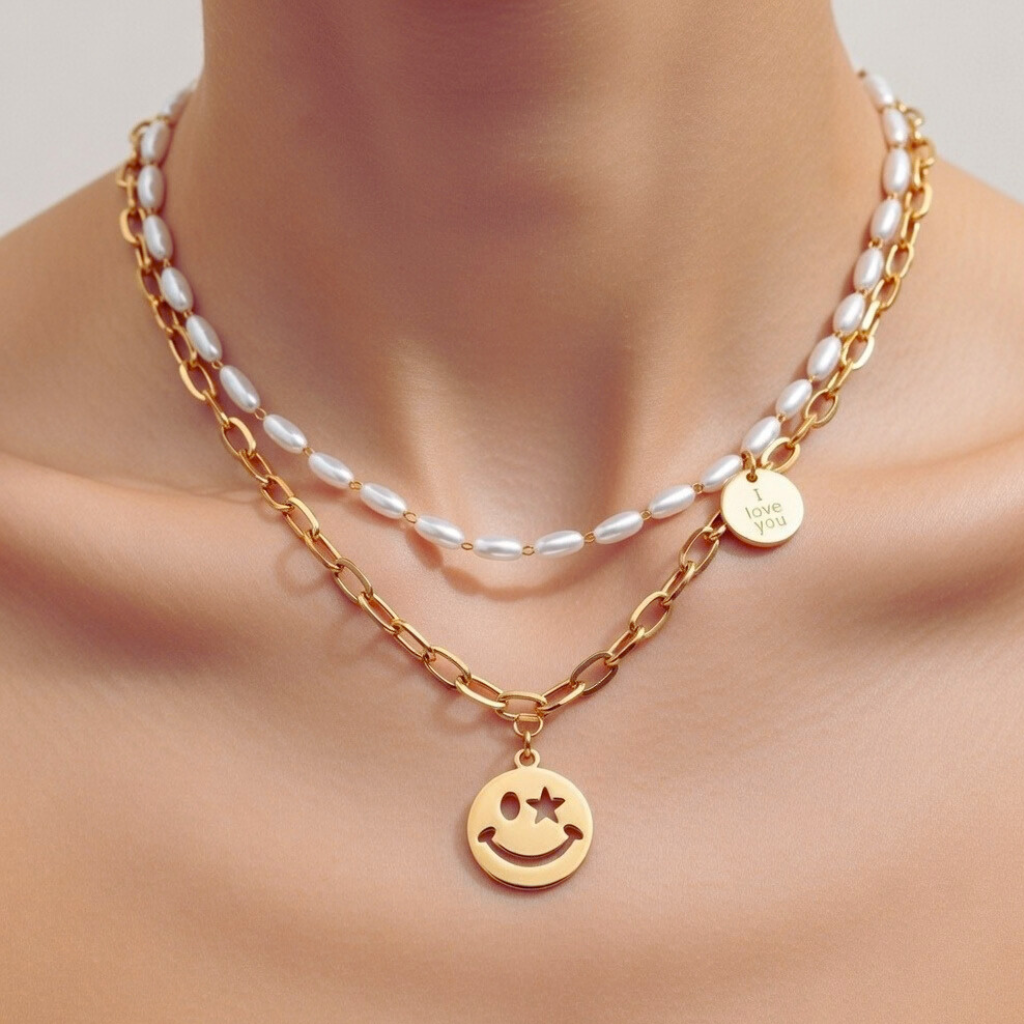 Nahaufnahme einer Frau, die ein zweireihiges goldenes Ketten-Set trägt. Die obere Kette besteht aus feinen, ovalen weißen Perlen mit einem kleinen runden Anhänger und der Gravur ‚I love you‘. Die untere, grobe Gliederkette trägt einen großen Smiley-Anhänger mit einem ausgestanzten Stern-Auge.