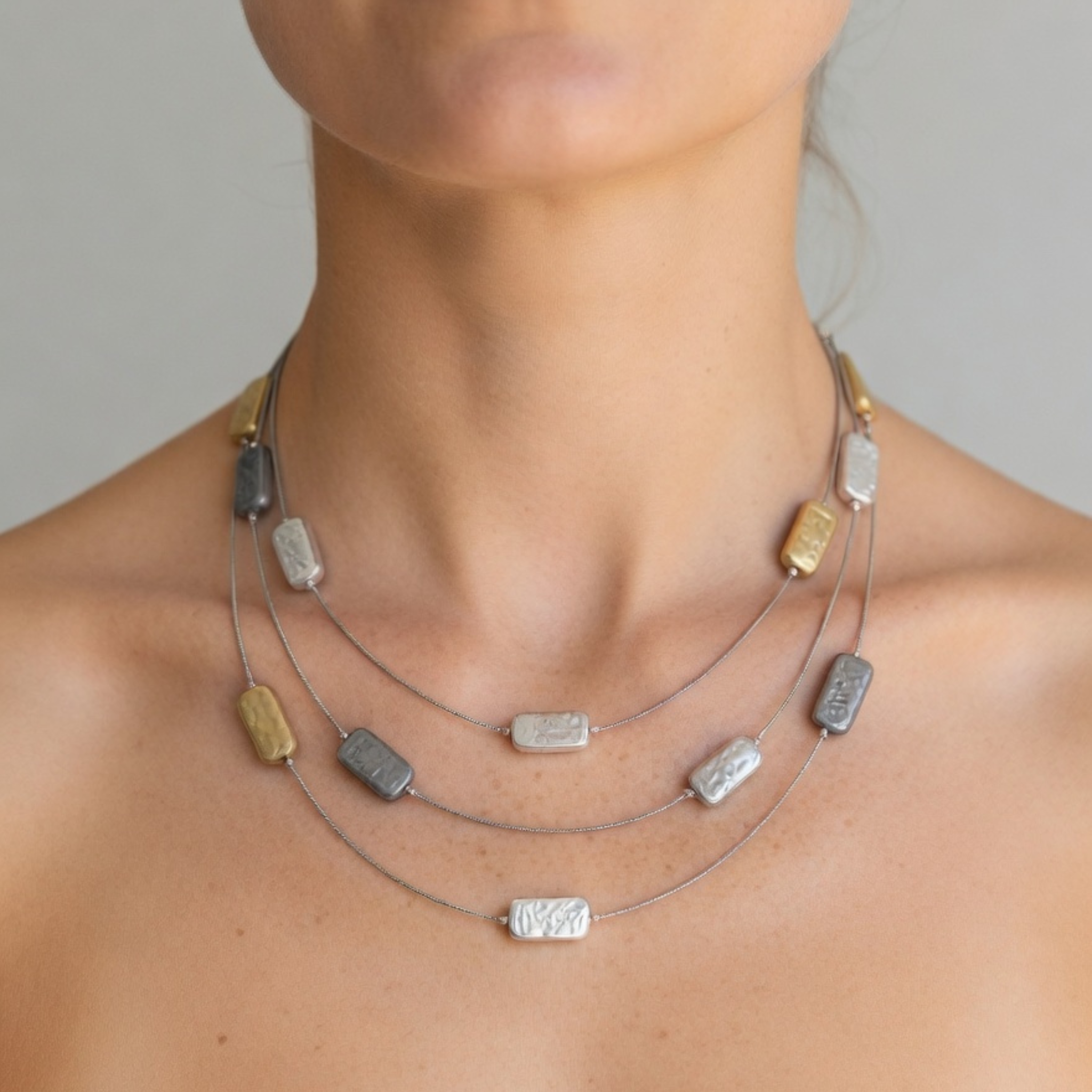 Nahaufnahme einer dreireihigen Collier-Halskette am Dekolleté einer Frau. Die feinen Kordeln tragen rechteckige, gehämmerte Design-Elemente in Silber, Grau und Gold.