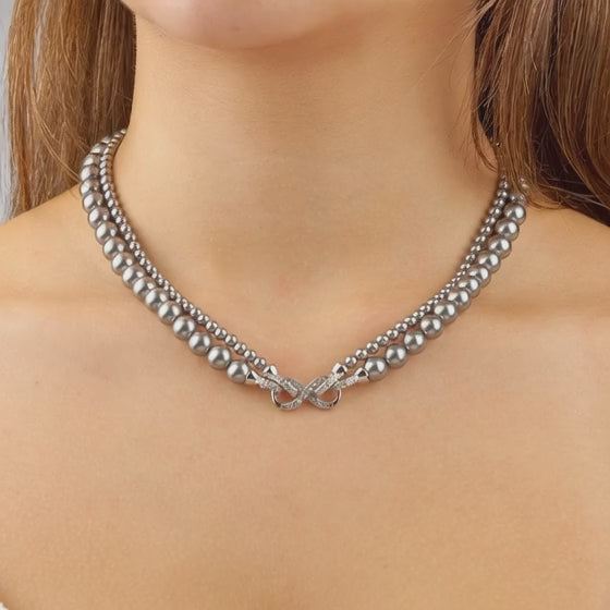 Eine Frau trägt ein elegantes, zweireihiges Collier aus anthrazitfarbenen Perlen. Im Zentrum liegt eine funkelnde, schleifenförmige Diamantschließe, die das Dekolleté dezent betont.