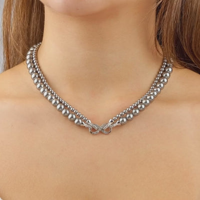 Eine Frau trägt ein elegantes, zweireihiges Collier aus anthrazitfarbenen Perlen. Im Zentrum liegt eine funkelnde, schleifenförmige Diamantschließe, die das Dekolleté dezent betont.